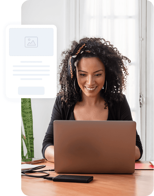 Woman with curly hair smiling while working on laptop at wooden desk with phone nearby, bright window in background.