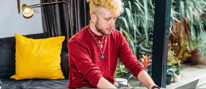 Man with blonde curly hair and red sweater working on a laptop indoors, with a yellow pillow and green plants nearby.