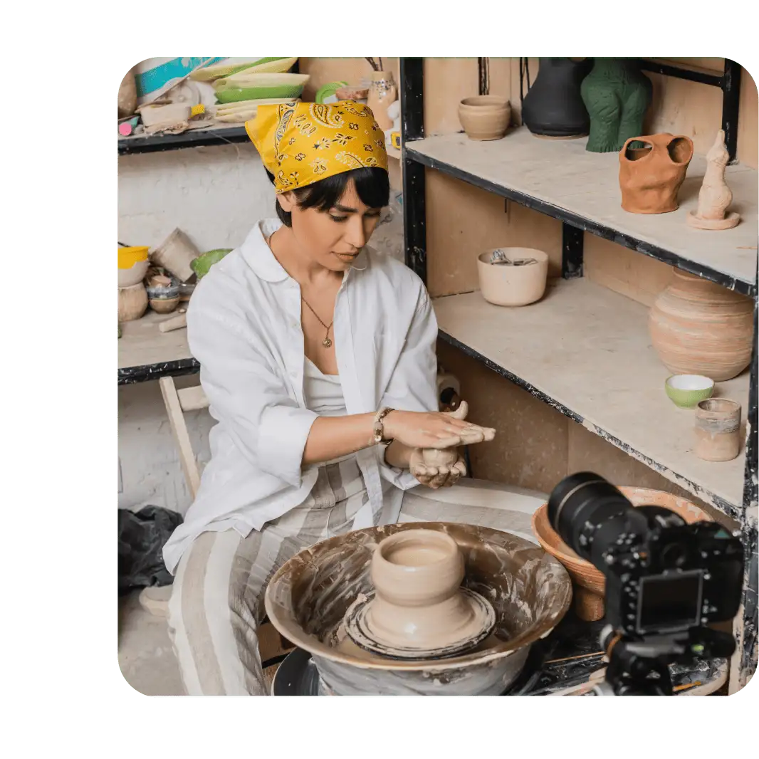 Woman in yellow bandana and white shirt shaping clay on a pottery wheel in a studio, filmed by a camera nearby.