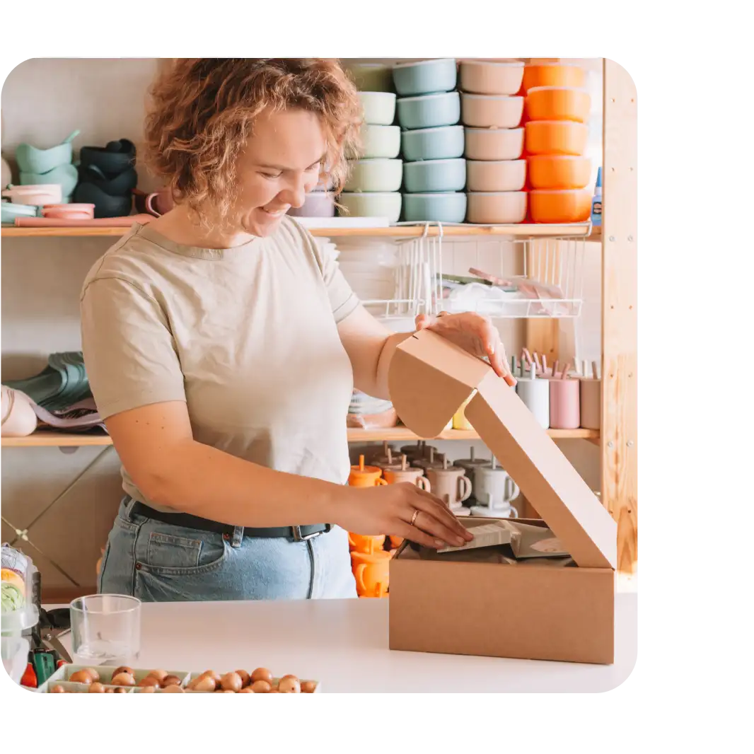A smiling woman opens a cardboard box in a colorful shop with shelves of stacked bowls and kitchenware behind her.