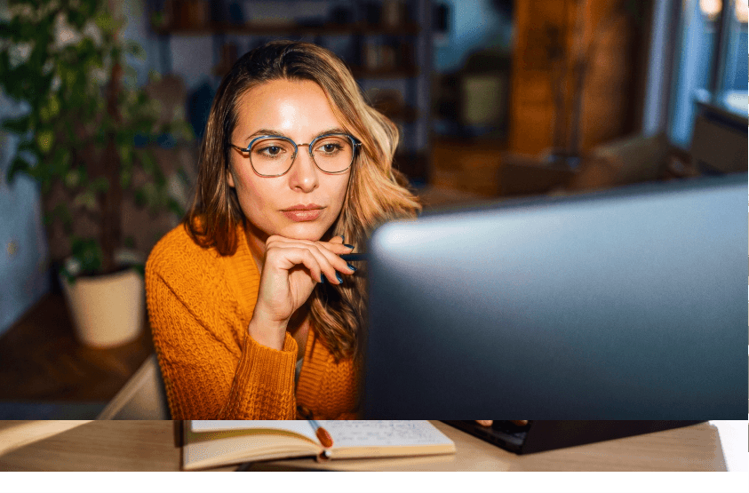 Woman with glasses looking thoughtfully at a computer screen in a home office setting.