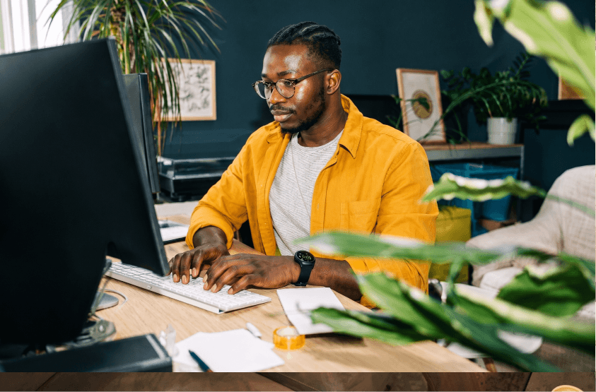 Man with glasses typing on a computer keyboard at a desk surrounded by indoor plants.