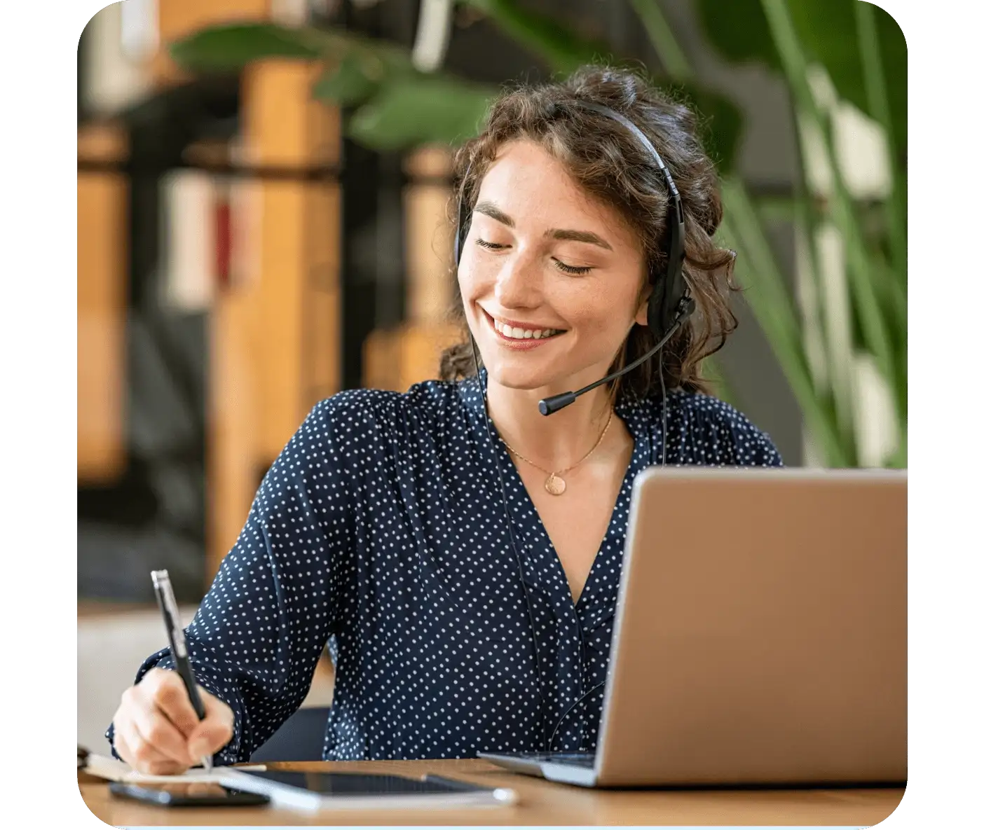Smiling customer service representative in blue polka dot blouse wearing headset while taking notes and using laptop.