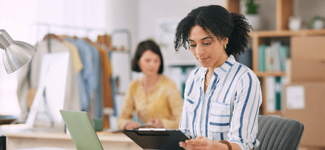 A woman sitting at a desk with a laptop and a clipboard in hand