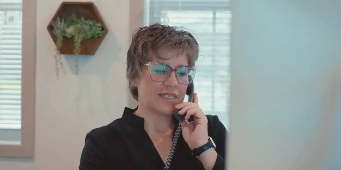 Person in blue glasses talking on phone in bright office with hexagonal plant holder on wall behind them.