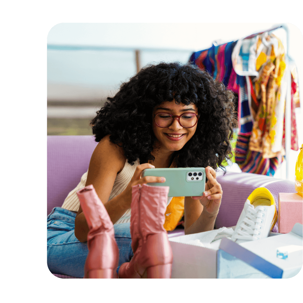 A smiling woman with curly hair and glasses photographs pink boots with her phone, surrounded by shoes and colorful clothes on a rack.