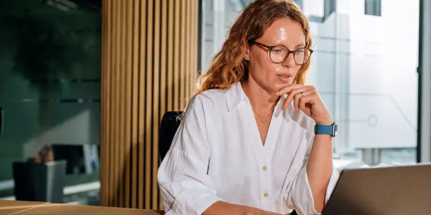 Woman with glasses and red hair in a white shirt thoughtfully working on a laptop in a modern office.