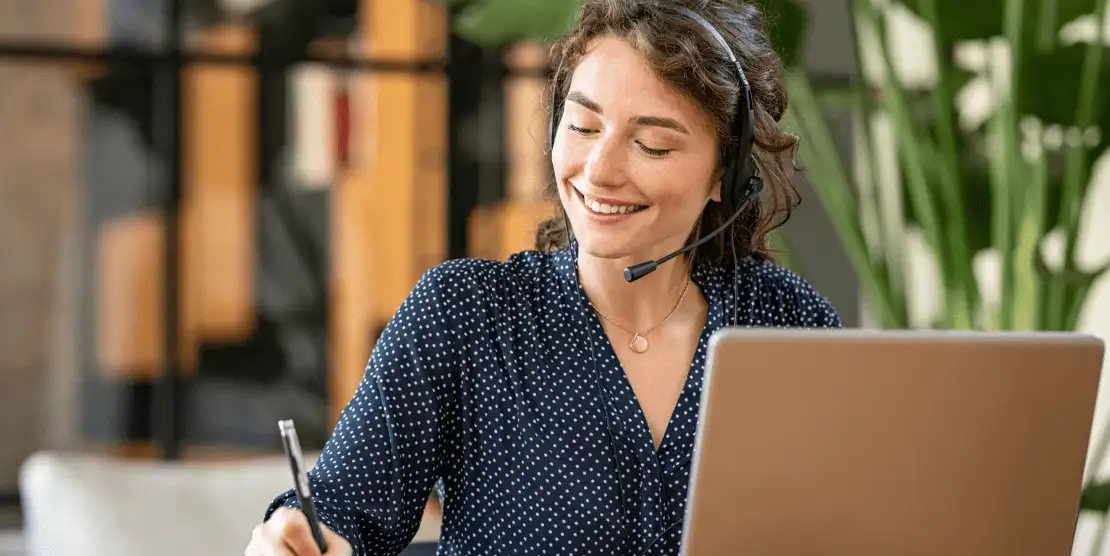 Woman in front of laptop with headset and writing on pad