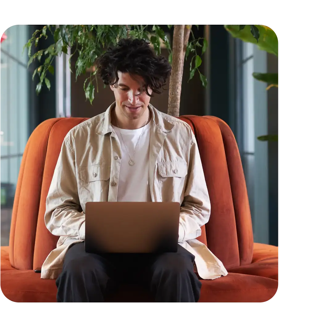 A young man with curly hair sits on an orange sofa, smiling while using a laptop, with green plants in the background.
