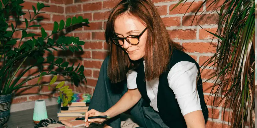 A woman with glasses and red hair writes in a notebook near plants and books against a brick wall.