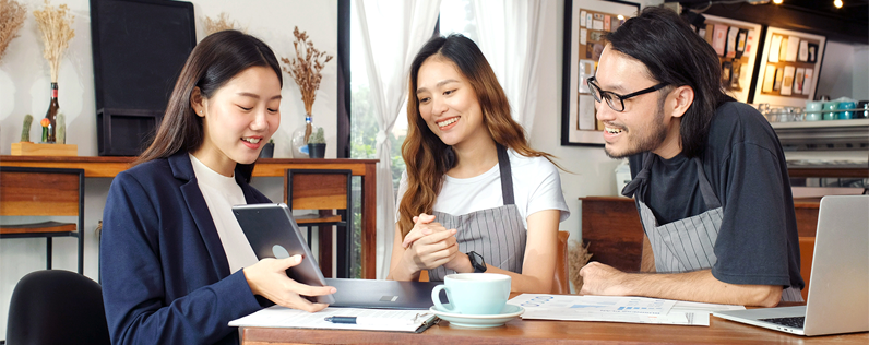 Header_HowCustomersFindBusiness Three people sit at a table in a coffee shop and discuss business plans. One holds a tablet and there are documents on the table.