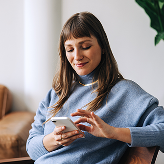 A smiling woman seated in a chair looks down at her smartphone.