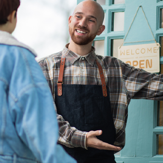 A man wearing an apron holds the door of a cafe open for a customer. The door is bright blue and has an open sign hanging from the top.
