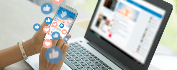 A hand holding a smartphone with emojis flying out of it in front of a laptop.