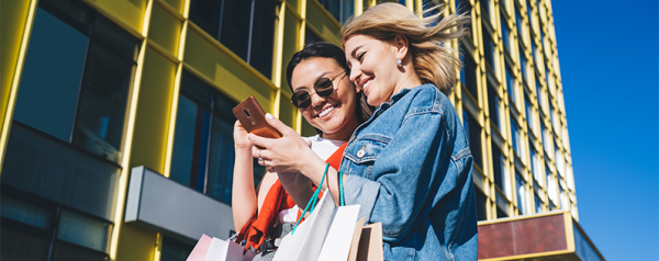 Two women stand on a city sidewalk and scroll on a smartphone. They each hold multiple colorful shopping bags.
