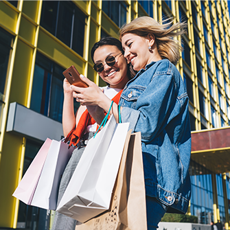 Two women stand on a city sidewalk and scroll on a smartphone. They each hold multiple colorful shopping bags.