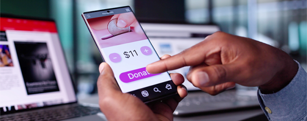 A person uses their mobile phone to donate to a fundraiser. A laptop is in the background with the screen showing an email.