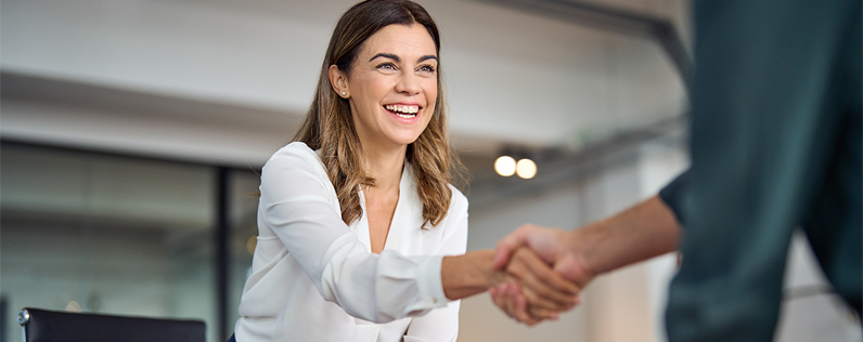 Header_CanadianBusinessServices A woman stands and leans over a desk to shake a partner's hand to solidify a business services agreement.