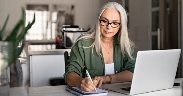 A woman sitting at a desk with her laptop, writing in a notebook