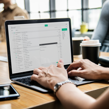 Hands type on a laptop placed on a conference room table with a coffee cup in the background.