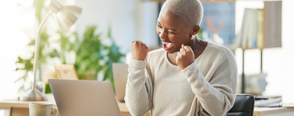 A woman sits at her desk and lifts her fists in a cheer. She smiles down at her laptop and gives a celebratory shout.