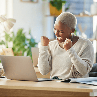 A woman sits at her desk and lifts her fists in a cheer. She smiles down at her laptop and gives a celebratory shout.