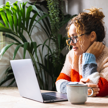 A woman sits in front of a laptop with a mug next to her.