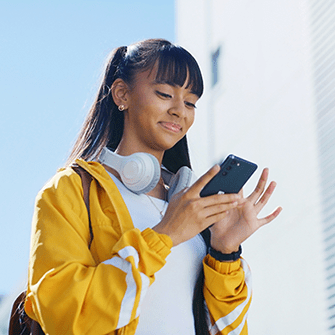 A woman in a yellow jacket with headphones around her neck looks down at her smartphone.