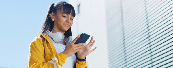 A woman in a yellow jacket with headphones around her neck looks down at her smartphone.