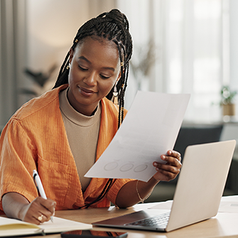 A woman sits at a desk in front of a laptop holding papers and writing with a pen.