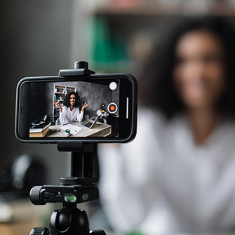 A woman sits behind a desk filming herself on her iPhone.