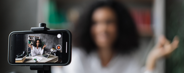 A woman sits behind a desk filming herself on her iPhone.