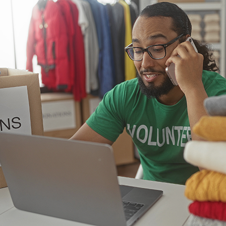 A man in a green "Volunteer" shirt talks on the phone while sitting in front of a laptop.