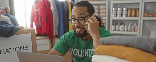 A man in a green "Volunteer" shirt talks on the phone while sitting in front of a laptop.