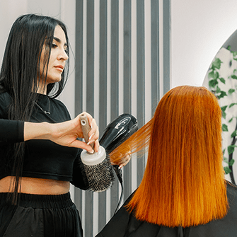 A hairstylist blowdries a client's hair in front of a mirror.