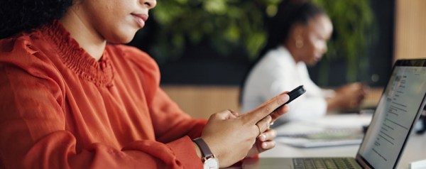 An image of a woman in front of a laptop checking her smartphone. In the blurred background, a colleague works at another desk in a modern office.