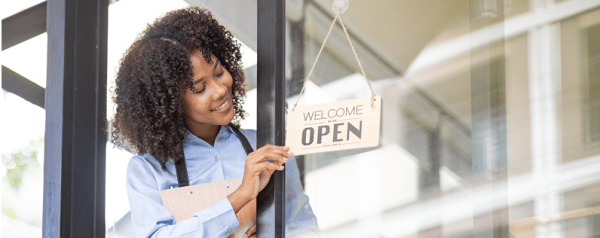 A woman wearing an apron and holding a clipboard hangs a wooden open sign on a glass door.