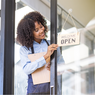 A woman wearing an apron and holding a clipboard hangs a wooden open sign on a glass door.