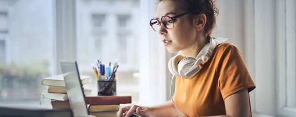 A young woman with glasses and headphones around her neck focusing on a laptop at a desk with books and pens.