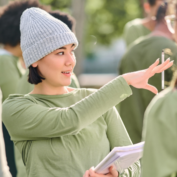 A woman in a beanie holds a stack of papers and a pen.