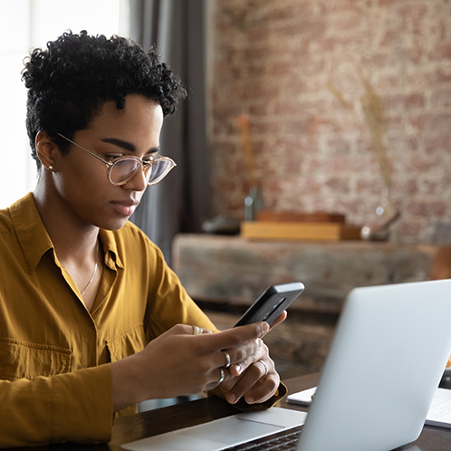 A person sits in front of a laptop looking at their smartphone