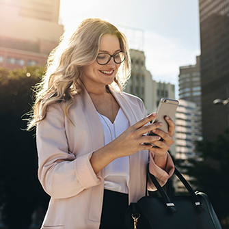 A woman standing in front of tall buildings smiles while looking down at her phone.