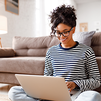 A woman in glasses sits on the floor looking at her laptop.
