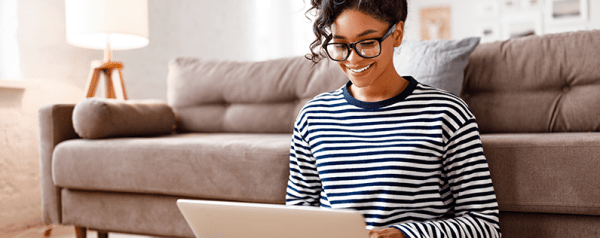A woman in glasses sits on the floor looking at her laptop.