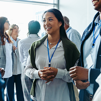 An image of a woman wearing a lanyard networking with a group of professionals at a conference event. In the background, other attendees stand in small groups chatting