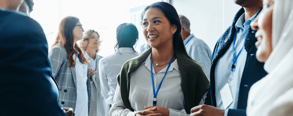 An image of a woman wearing a lanyard networking with a group of professionals at a conference event. In the background, other attendees stand in small groups chatting