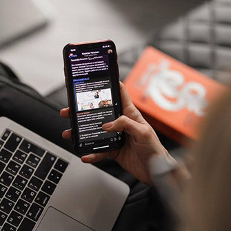 A hand scrolls on a smartphone with a laptop in the background