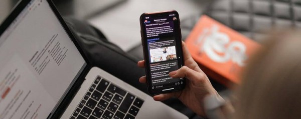 A hand scrolls on a smartphone with a laptop in the background