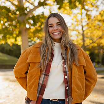A smiling woman in an orange jacket stands in front of trees with fall leaves.