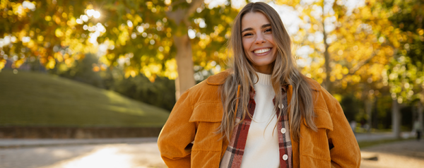 A smiling woman in an orange jacket stands in front of trees with fall leaves.